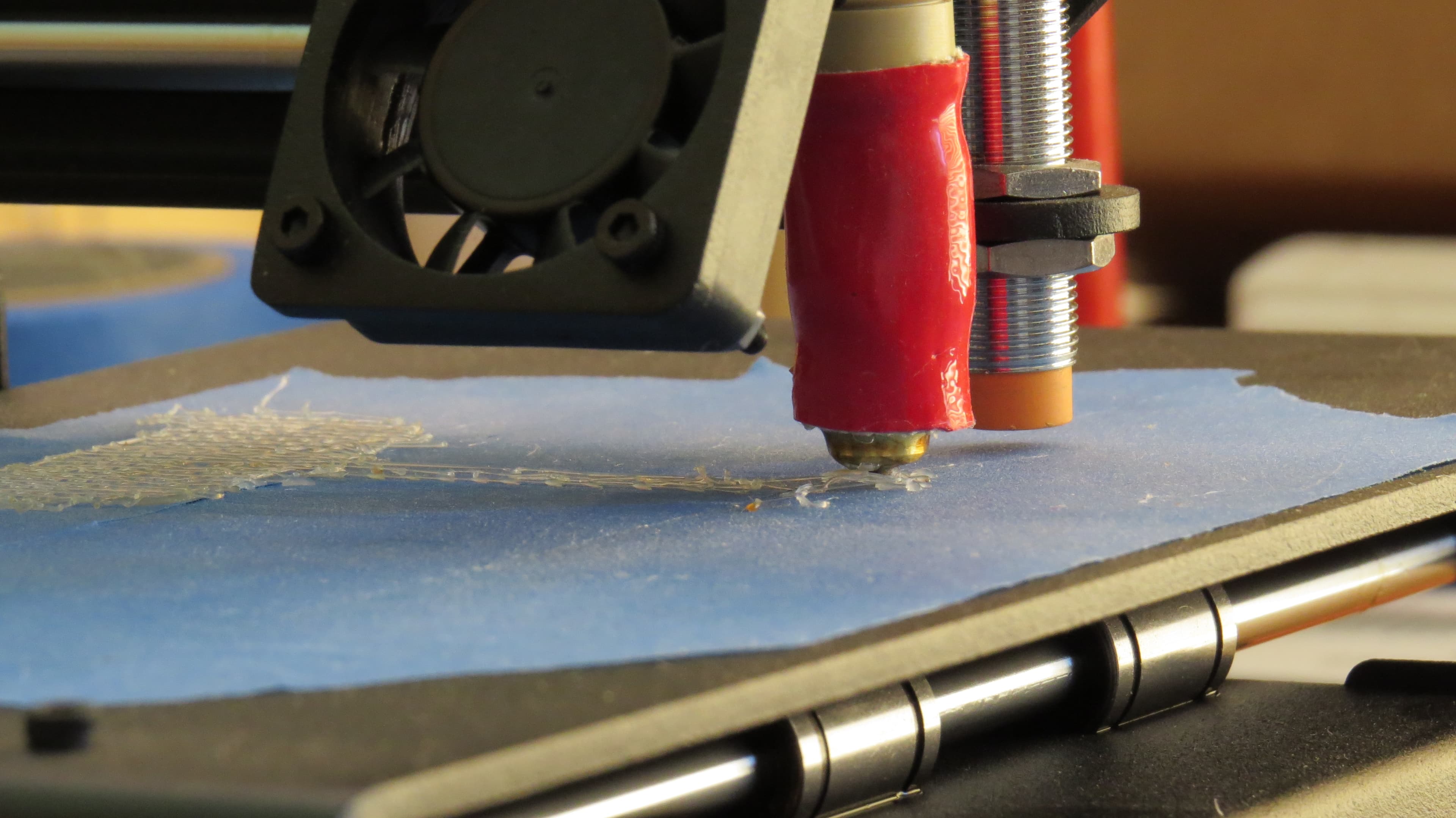 Fotografía de primer plano de una boquilla FDM depositando la primera capa sobre cinta azul en la cama de impresión